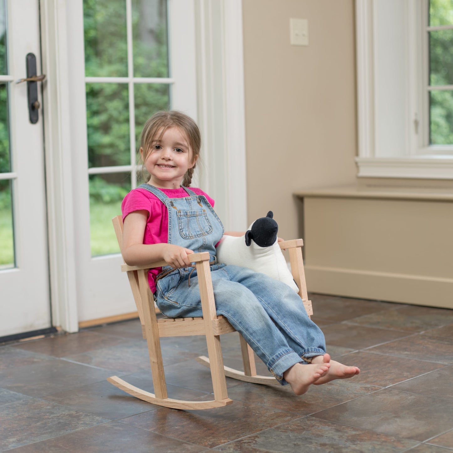 Young child sitting happily on a handcrafted Amish made wooden rocker made of unfinished ash, holding a black and white plush toy, set indoors by glass doors, part of Toys, Books & Games and USA Made collections.