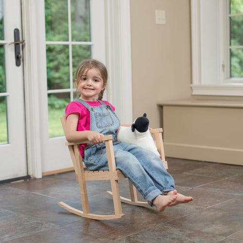 Young child sitting happily on a handcrafted Amish made wooden rocker made of unfinished ash, holding a black and white plush toy, set indoors by glass doors, part of Toys, Books & Games and USA Made collections.