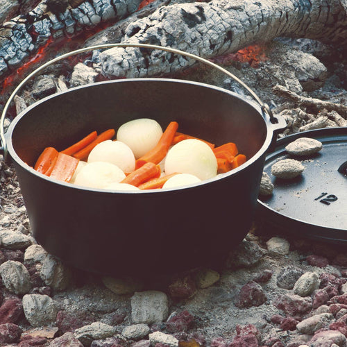 Handcrafted cast iron camp Dutch oven with built-in legs cooking whole onions and carrot pieces over hot coals in an outdoor campfire setting; featured in Farm, Garden & Tool, Camping, Cast Iron, USA Made, Best Sellers, and Promo Eligible collections.