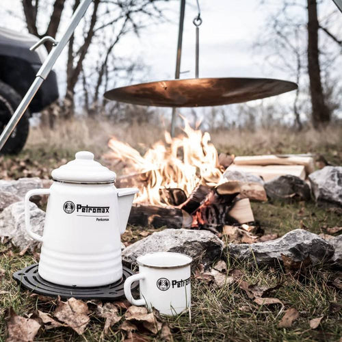 White enamel Petromax coffee pot and matching mug with black logo, placed on a black trivet near a campfire outdoors, perfect for Camping, Kitchen & Food, and Brewing Coffee and Tea collections.