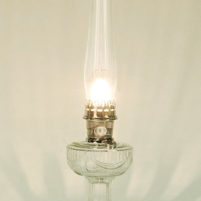 Lit vintage-style clear glass oil lamp with a tall chimney and metal burner, featured in Lamps & Lighting, Parts and Accessories, Wicks and Mantles, Lamp Burners and Collars, Best Sellers, and Promo Eligible collections.