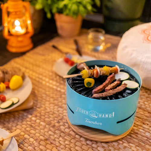 Handcrafted blue tabletop charcoal grill cooking skewers and sliced vegetables, set on a woven mat near a glowing lantern, ideal for Farm, Garden & Tool, Camping, and Grilling collections.