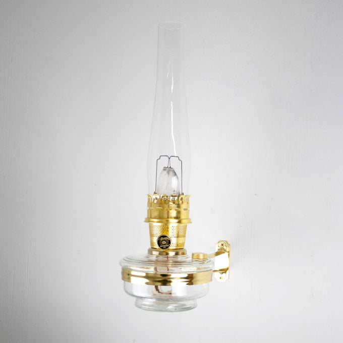 Handcrafted brass wall bracket with clear glass oil lamp and tall chimney, designed for classic Aladdin lamps, featured in Lamps & Lighting, Oil Lamps, Wall Lamps, Best Sellers, and Promo Eligible collections.