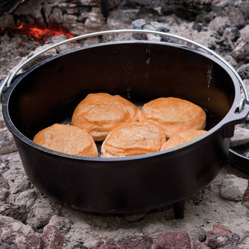 Handcrafted Lodge cast iron camp Dutch oven with built-in legs and metal bail handle baking golden biscuits over glowing coals; featured in Farm, Garden & Tool, Camping, Cast Iron, USA Made, Best Sellers, Promo Eligible collections.