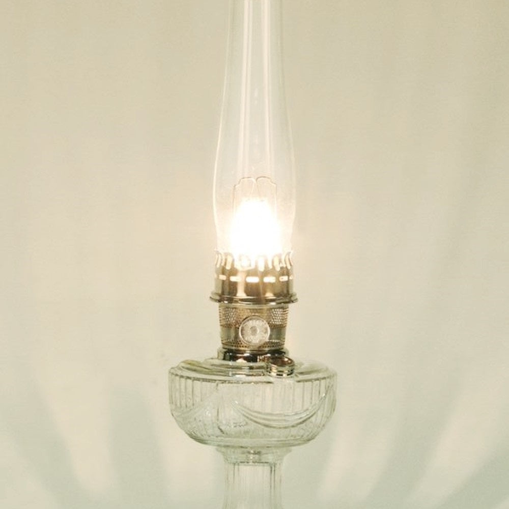 Lit clear glass oil lamp with a tall chimney and metal burner, emitting a warm glow, featured in Lamps & Lighting, Parts and Accessories, Wicks and Mantles, Lamp Burners and Collars, Best Sellers, and Promo Eligible collections.