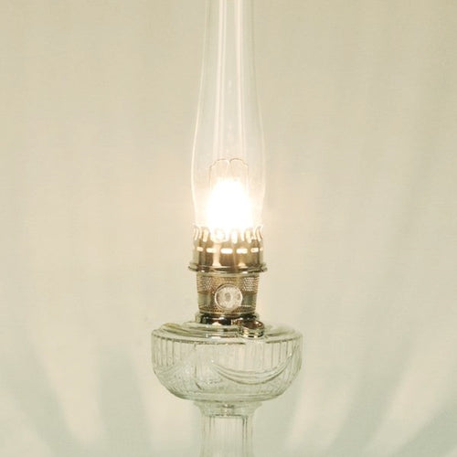 Lit clear glass oil lamp with a tall chimney and metal burner, emitting a warm glow, featured in Lamps & Lighting, Parts and Accessories, Wicks and Mantles, Lamp Burners and Collars, Best Sellers, and Promo Eligible collections.