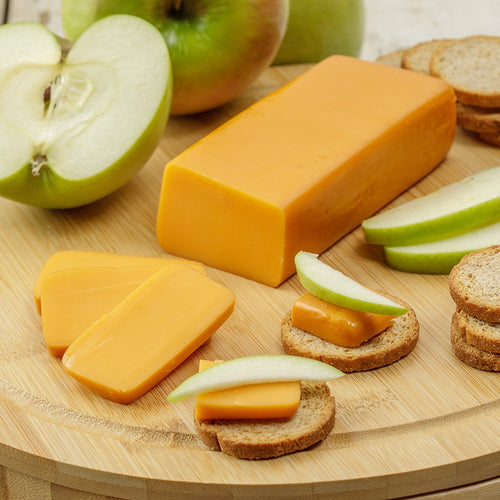Block of handcrafted shelf-stable cheese with smooth orange color, sliced and served on oat crackers with green apple wedges, ideal for Kitchen & Food, Pantry, Snacks and Sweets, USA Made, and Best Sellers.