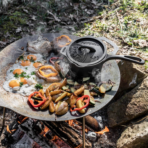 Handcrafted cast iron saucepan with lid cooking over an open fire on a steel tripod, surrounded by fresh eggs, roasted potatoes, mushrooms, and bell peppers, ideal for farm, garden, and camping cookware collections.