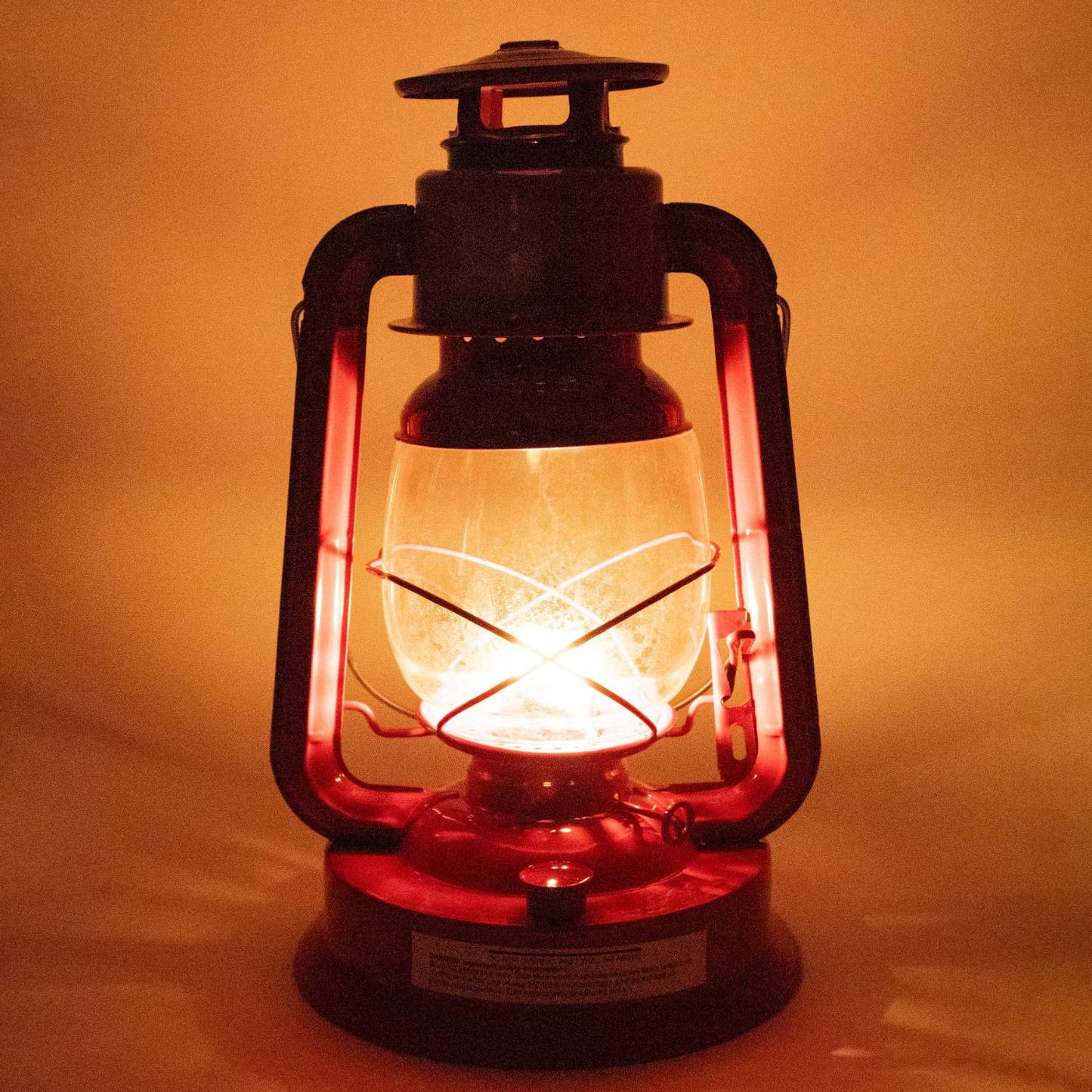 Glowing red metal oil lantern with glass globe casting warm light, ideal for outdoor use in camping, pest control, emergency supplies, and farm or garden settings.