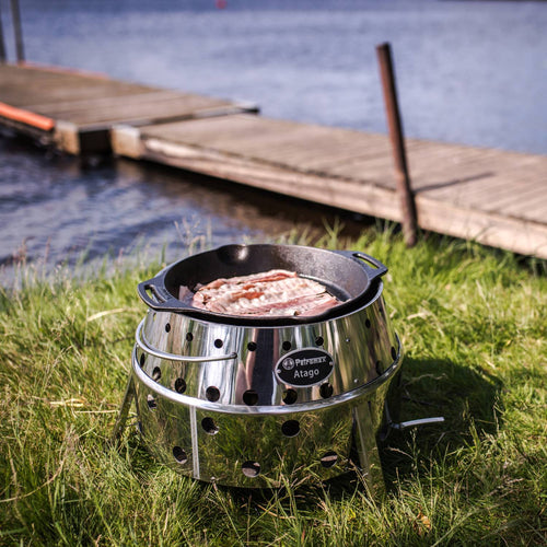 Portable stainless steel camp grill and fire bowl with ventilation holes and fold-out legs, shown cooking bacon outdoors by a lake, ideal for camping and grilling collections.