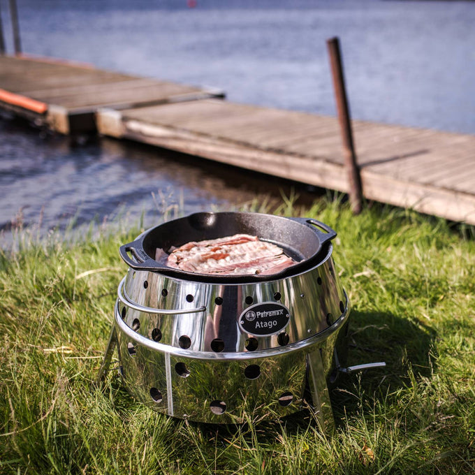 Portable stainless steel camp grill and fire bowl with ventilation holes and fold-out legs, shown cooking bacon outdoors by a lake, ideal for camping and grilling collections.