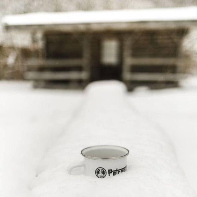White enameled steel mug with metal rim and black logo, resting on a snow-covered surface in front of a rustic wooden cabin, ideal for Farm, Garden & Tool, Camping, and Brewing Coffee and Tea collections.