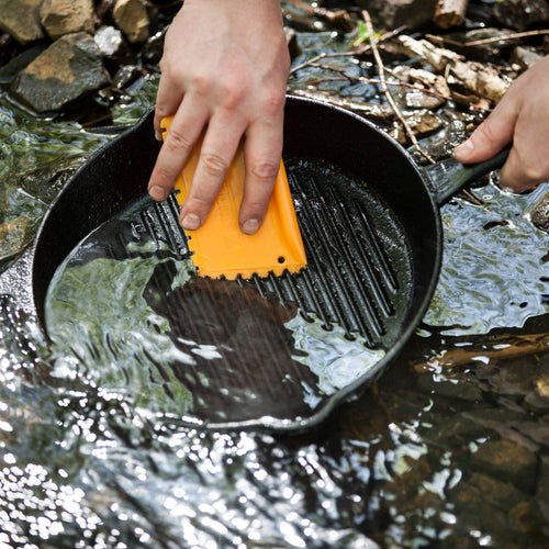 Hands using a bright orange pan scraper to clean a cast iron grill pan under flowing water outdoors, highlighting durable cookware care in Kitchen & Food, Cookware, Cast Iron, Best Sellers, and Promo Eligible collections.