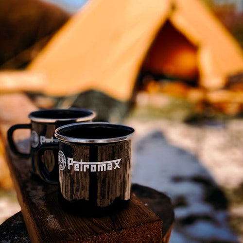 Two black enamel steel mugs with white logo and metal rims resting on a wooden surface outdoors near a tan canvas tent, ideal for Camping, Farm, Garden & Tool, and Brewing Coffee and Tea collections