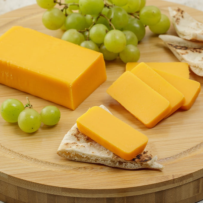 Block of handcrafted shelf-stable cheese sliced and placed on a piece of flatbread, accompanied by green grapes on a round wooden board, ideal for Kitchen & Food, Pantry, and USA Made collections.