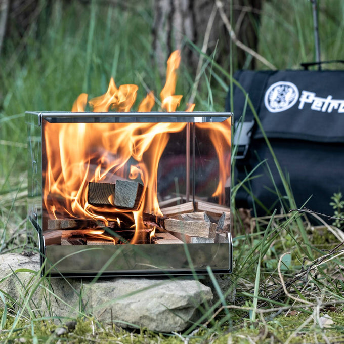 Compact handcrafted stainless steel camping stove burning wood in a natural outdoor setting with flames visible, accompanied by a branded carrying case, ideal for Farm, Garden & Tool, Camping, Emergency Supplies, Best Sellers, and Promo Eligible collections.