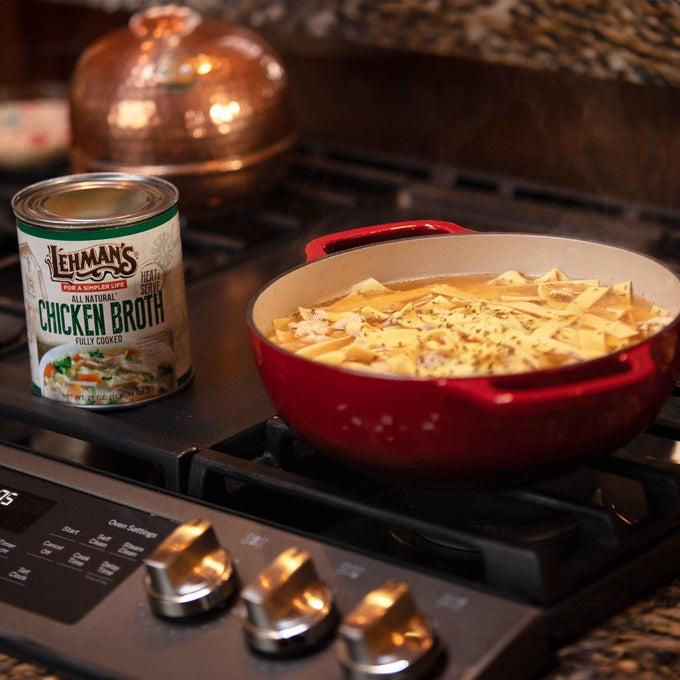 Lehman's canned chicken broth displayed on a stove next to a red pot simmering noodle soup, highlighting farm, kitchen, and emergency food supplies in a warm kitchen setting
