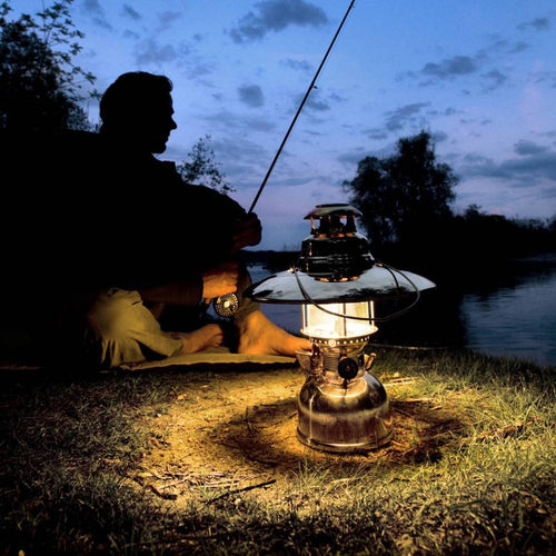 Glowing vintage-style pressure lamp with a polished metal top reflector illuminating a peaceful lakeside scene where a silhouette of a person fishes at dusk, ideal for lamps and lighting accessories in best sellers and promo collections.