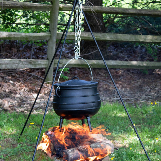 Unique cast iron campfire kettle suspended over an open wood fire by a tripod, designed for outdoor cooking in Farm, Garden & Tool, Camping, Best Sellers, and Promo Eligible collections.
