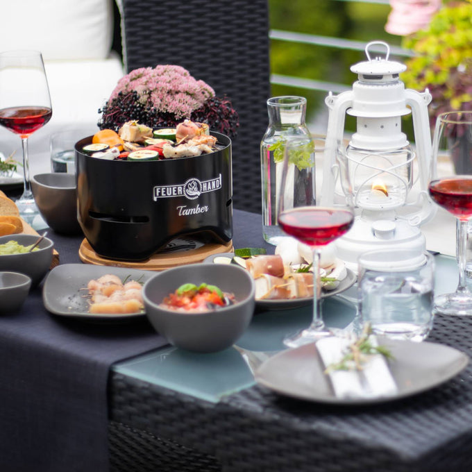 Black tabletop charcoal grill with food cooking on the grate, set on a wooden trivet amid an outdoor dining arrangement featuring wine glasses, a white lantern, and garden greenery, perfect for Farm, Garden & Tool and Grilling collections.