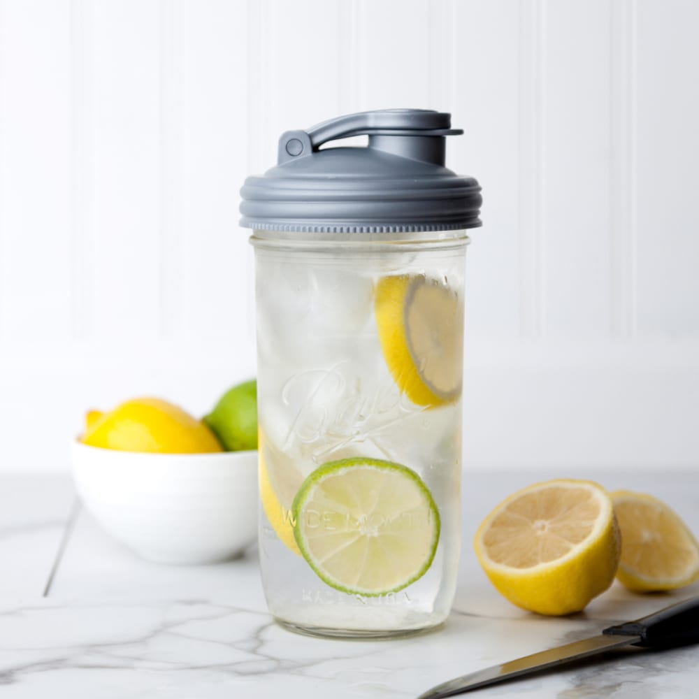 Clear wide-mouth mason jar filled with ice water and slices of lemon and lime, topped with a gray pour lid featuring a carry loop, shown on a marble countertop with fresh citrus in the background, perfect for Kitchen & Food, Dinnerware, and USA Made collections.