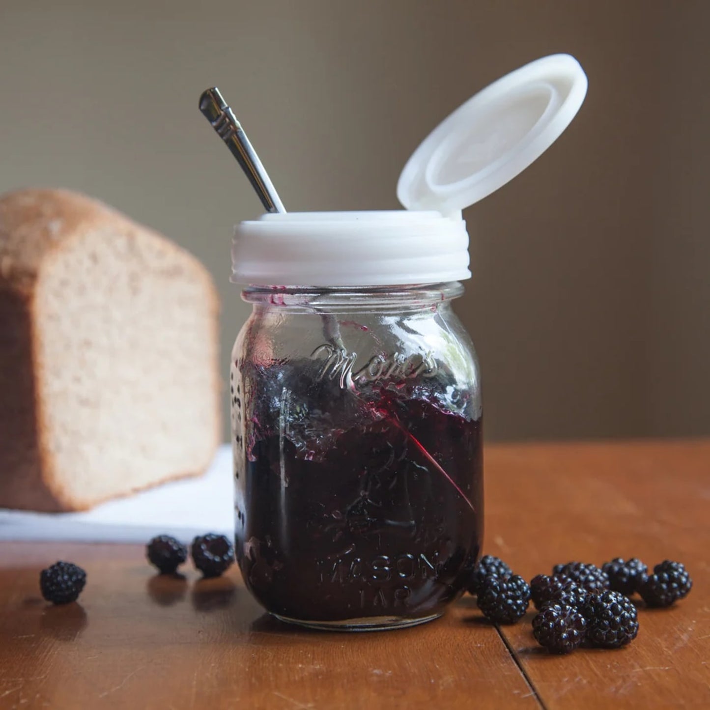 Glass Mason jar filled with blackberry jam, featuring a white flip top lid open to reveal a spoon inside, displayed with fresh blackberries and a loaf of bread in a kitchen setting, handcrafted and made in USA, suitable for Home & Decor, Kitchen & Food, and Storage and Serving collections.
