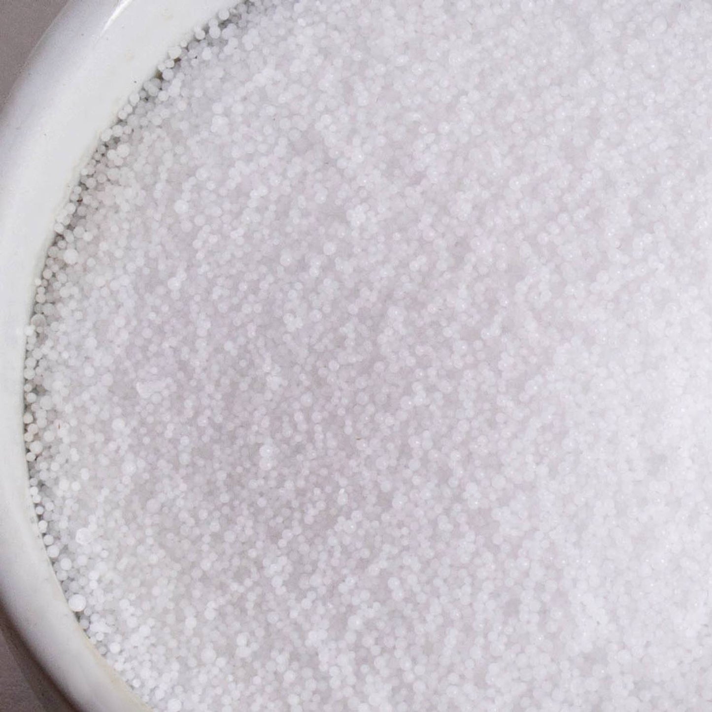 Close-up of white food-grade sodium hydroxide lye beads in a white ceramic container, used for soapmaking and featured in Health & Wellness and Soapmaking Supplies collections.