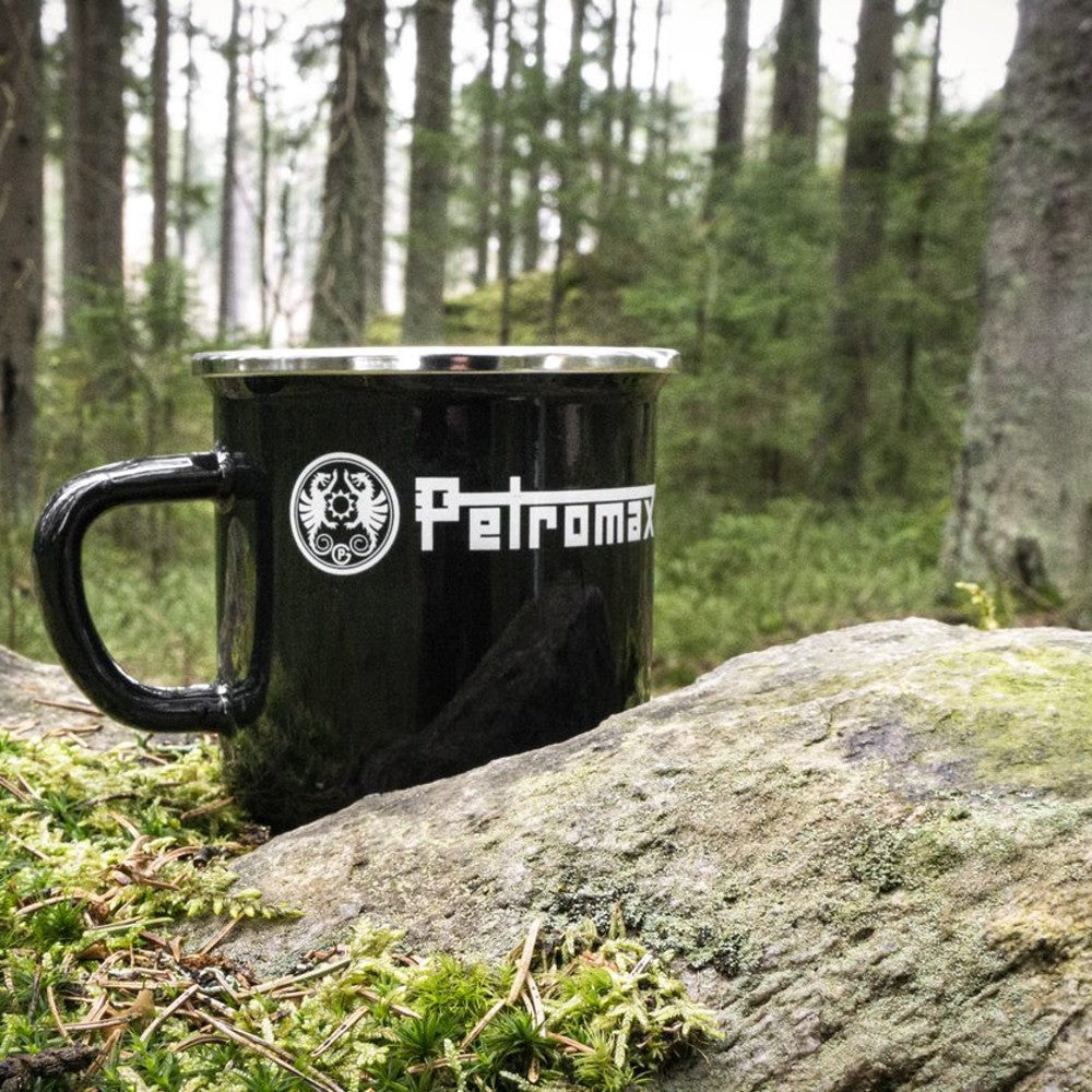 Black enameled steel camping mug with metal rim and bold logo, resting on mossy ground near a forest rock, ideal for Farm, Garden & Tool, Camping, and Brewing Coffee and Tea collections