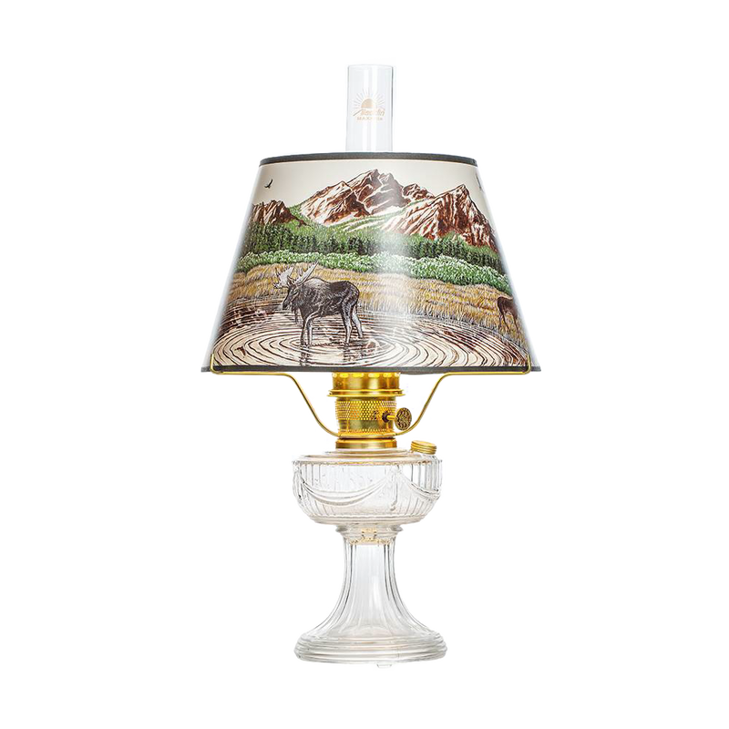 Clear oil lamp with brass burner and a Rocky Mountain scenic parchment shade depicting a moose by a lake with mountains in the background, featured in Lamps & Lighting, Oil Lamps, Table Lamps, Best Sellers, and Promo Eligible collections.