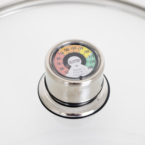 Close-up of a stainless steel built-in temperature gauge on a glass lid with color-coded heat zones, designed for precise monitoring in canning, food preservation, and kitchen use.