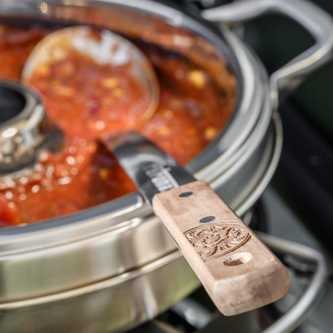 Handcrafted stainless steel serving spoon with a long walnut handle featuring an engraved logo, resting on a pot of hearty stew, suitable for Farm, Garden & Tool, Kitchen & Food, Camping, Storage and Serving, Best Sellers, Promo Eligible collections.