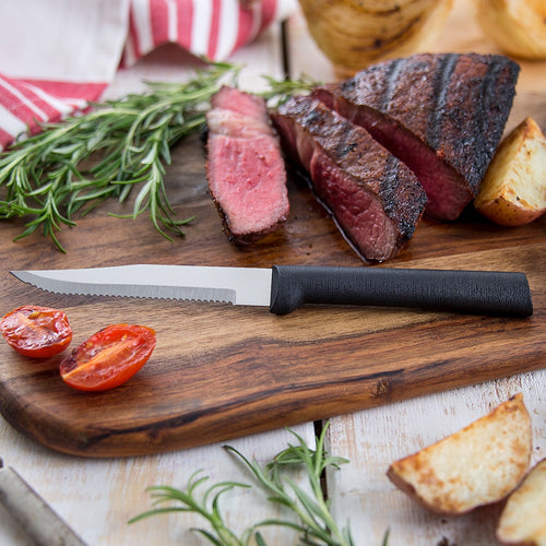 Handcrafted serrated steak knife with high carbon surgical stainless steel blade and solid black aluminum handle resting on a wooden board with sliced grilled steak, roasted potato, tomato, and fresh rosemary, ideal for Kitchen & Food, Farm, Garden & Tool, Knives & Axes, USA Made, and Best Sellers collections.