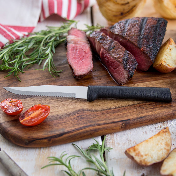 Handcrafted serrated steak knife with high carbon surgical stainless steel blade and solid black aluminum handle resting on a wooden board with sliced grilled steak, roasted potato, tomato, and fresh rosemary, ideal for Kitchen & Food, Farm, Garden & Tool, Knives & Axes, USA Made, and Best Sellers collections.