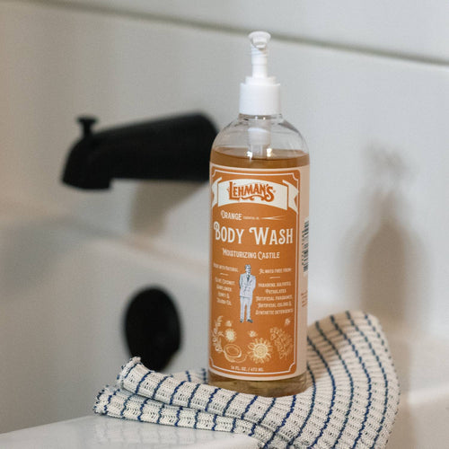 Handcrafted moisturizing castile body wash in an amber-orange shade with pump dispenser, featuring natural plant-based oils, placed on a striped cloth by a bathtub faucet, part of USA Made, Health & Wellness, Grooming and Personal Hygiene, and Best Sellers collections.
