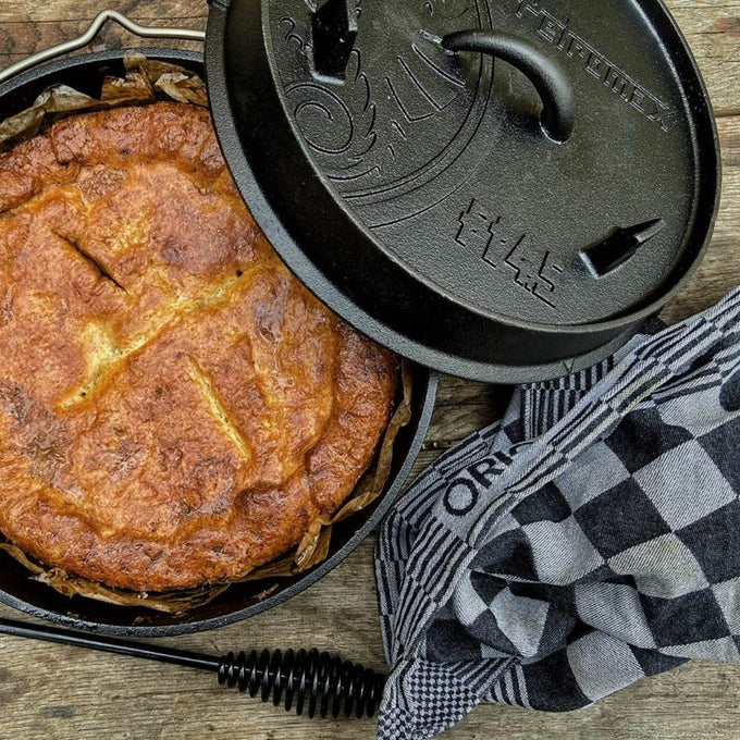 Cast iron Dutch oven with lid removed revealing a baked pie inside, placed next to a black and white checkered kitchen towel, suitable for outdoor and indoor cooking in Kitchen & Food and Cast Iron collections.