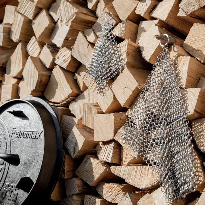Two sizes of stainless steel Petromax chainmail cleaners hanging on stacked firewood next to a cast iron lid, handcrafted for tough cleaning, featured in multiple home and cleaning collections.