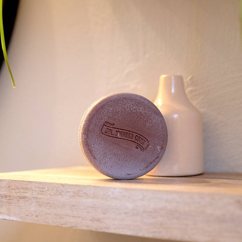 Round brown shaving soap bar with embossed branding, placed on a wooden shelf next to a simple beige ceramic vase, featured in Health & Wellness, Grooming and Personal Hygiene, Soaps and Soapmaking, USA Made, Best Sellers, and Promo Eligible collections.