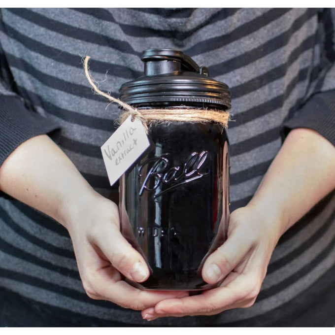 Hands holding a wide-mouth glass jar filled with dark vanilla extract, sealed with a black pour lid featuring a carry loop, suitable for Kitchen & Food, Dinnerware, and Kitchen Gadgets collections.