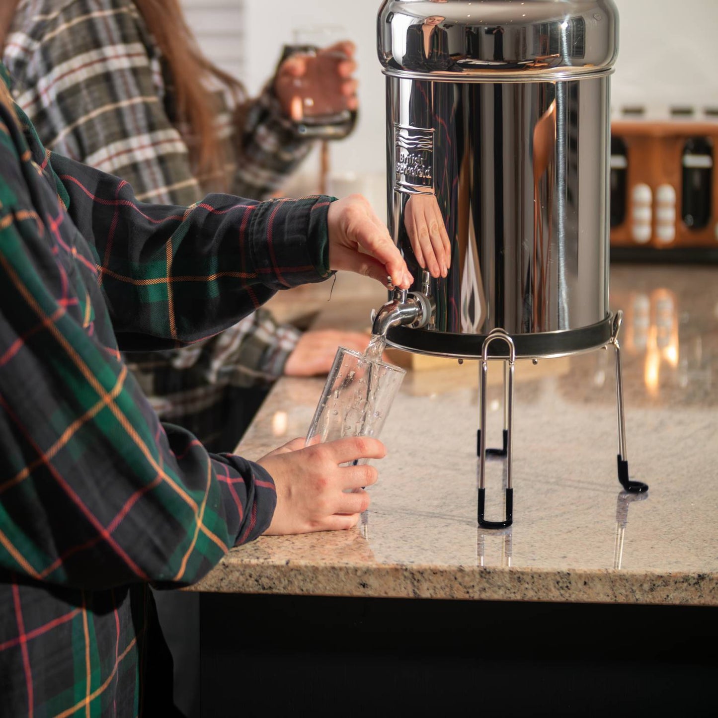 Person pouring water from a polished stainless steel gravity water filter elevated on a sturdy stand with protective feet, featured in Appliances, Emergency Supplies, and Best Sellers collections.