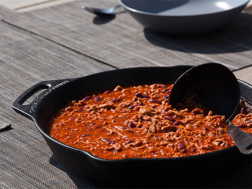 Cast iron skillet with long handle filled with seasoned chili containing beans and ground meat on a woven placemat, ideal for Farm, Garden & Tool, Kitchen & Food, Camping, Cookware, Cast Iron, and Best Sellers collections.