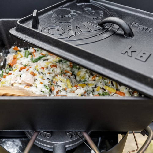 Close-up of a durable cast iron loaf pan with embossed lid cooking a hearty vegetable and rice casserole over a portable camp stove, ideal for camping, kitchen, and farm collections.