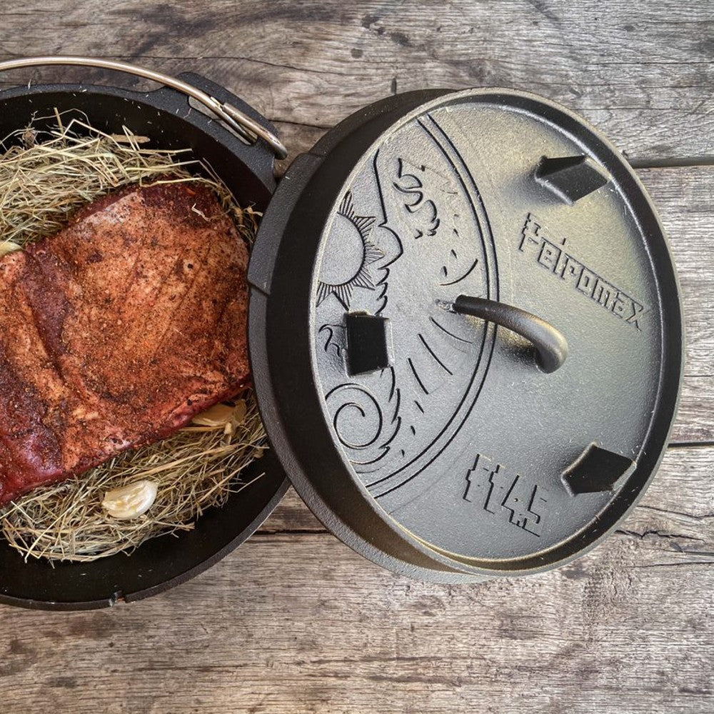 Open cast iron Dutch oven with legs, showing cooked seasoned meat resting on hay and garlic, featuring an embossed lid with decorative sun motif for Kitchen & Food, Cookware, Cast Iron collections.