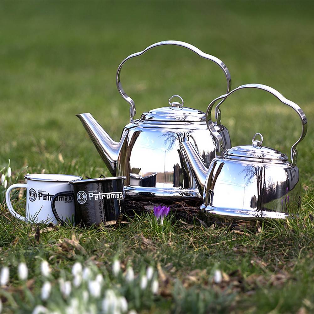 Two polished stainless steel teakettles with bent handles and secure lids sitting on grass surrounded by early spring flowers, accompanied by three enamel Petromax mugs, ideal for Farm, Garden & Tool, Kitchen & Food, Camping, Brewing Coffee and Tea, Best Sellers, and Promo Eligible collections.