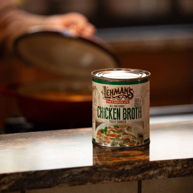 27 oz can of Lehman's all natural, fully cooked chicken broth placed on a kitchen counter with a pot lid being lifted in the background, ideal for Farm, Kitchen & Food, Emergency Supplies, Best Sellers, and Promo Eligible collections.