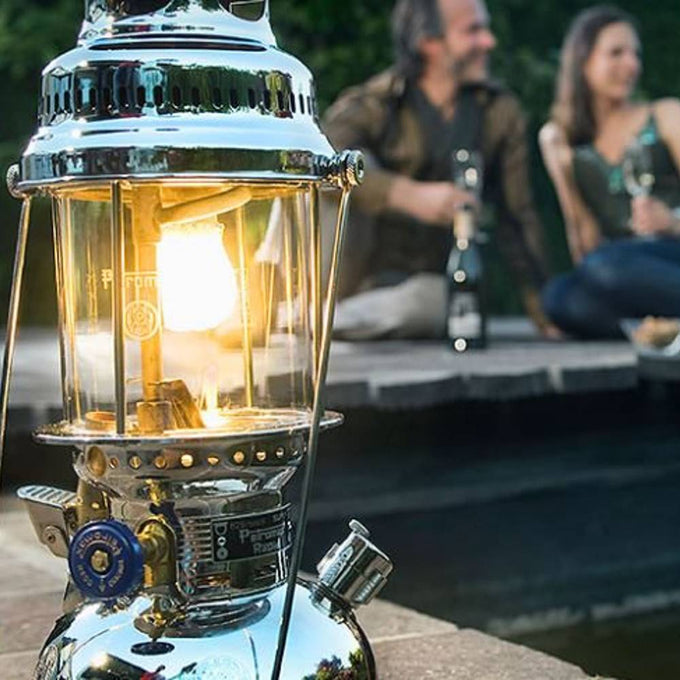 Close-up of a shining metal kerosene pressure lamp with a glowing mantle, surrounded by a blurred outdoor setting featuring two people relaxing, ideal for camping and emergency lighting.