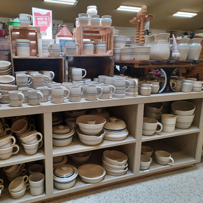 Handcrafted cream-colored stoneware kitchenware including mugs, crocks, and bowls with blue accent lines, displayed on shelves in a rustic food preservation and fermenting shop.