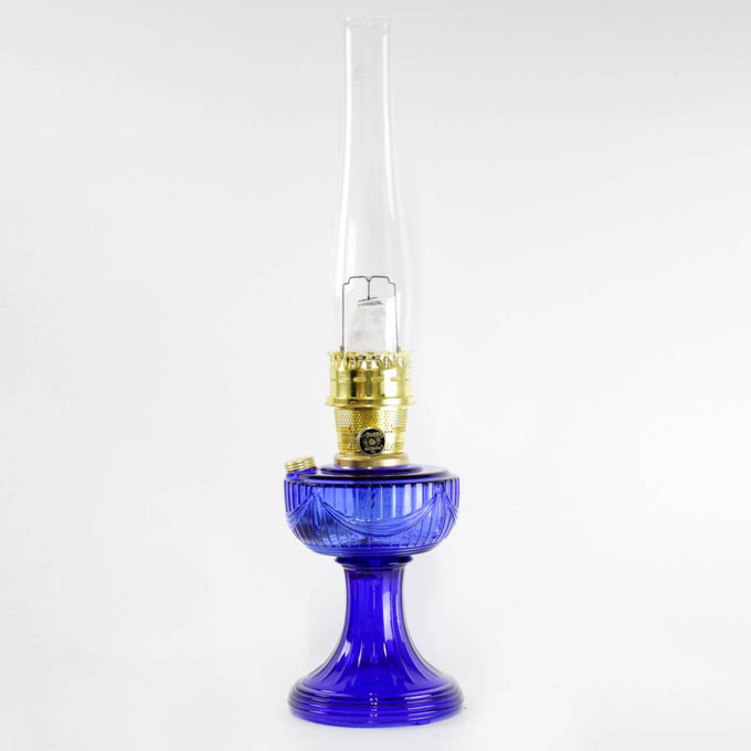 Handcrafted cobalt glass oil lamp with brass burner and tall clear chimney, featuring a Lincoln drape pattern, ideal for Lamps & Lighting, Oil Lamps, Table Lamps, Best Sellers, and Promo Eligible collections.