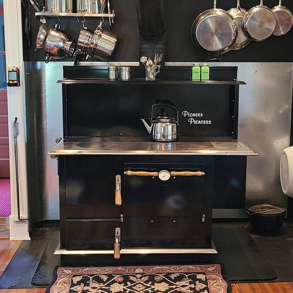 Black vintage-style cookstove with porcelain finish and wood handles, equipped with a kettle on top, surrounded by hanging metal pots and pans, ideal for Stoves, Cookstoves, and Best Sellers collections.