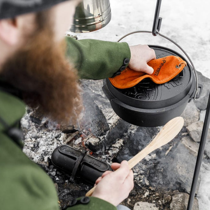 Man lifting lid off a sturdy cast iron Dutch oven with legs using an orange heat-resistant glove over an open campfire, surrounded by cookware and wooden utensils, ideal for Kitchen & Food, Cookware, Cast Iron, Best Sellers, and Promo Eligible collections.