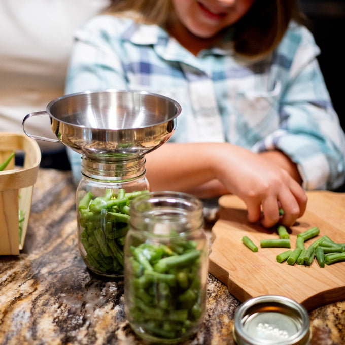 Child carefully packing fresh green beans into glass jars using a polished stainless steel funnel, showcasing a handcrafted canning process in Kitchen & Food, Food Preservation, and Canning collections.